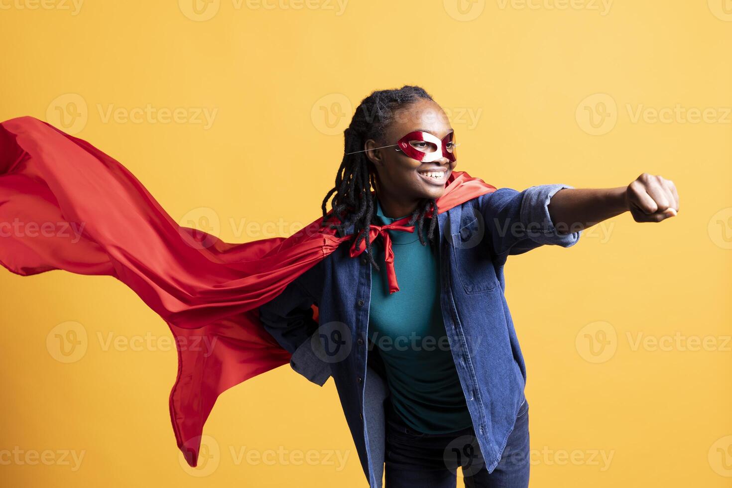 Happy girl portraying superhero flying with cape fluttering in wind, hiding identity, studio background. Upbeat woman posing as hero in costume, covering face with mask, acting enigmatic photo
