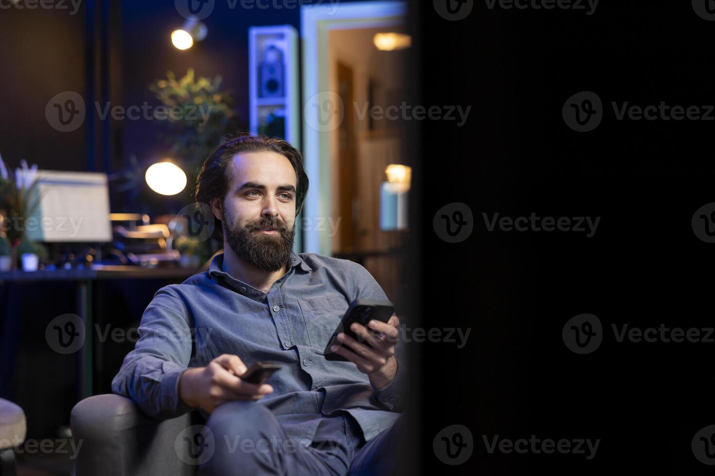 Smiling person on couch binging series on streaming service, browsing using remote and texting on mobile phone. Happy man watching shows on television, zapping though channels, chatting on cellphone photo
