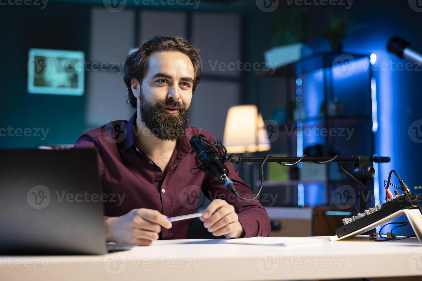 Happy influencer sitting at coffee table with microphone and mini house plants in dimly lit home studio interior, chatting with fans. Smiling man surrounded by podcast recording equipment in apartment photo