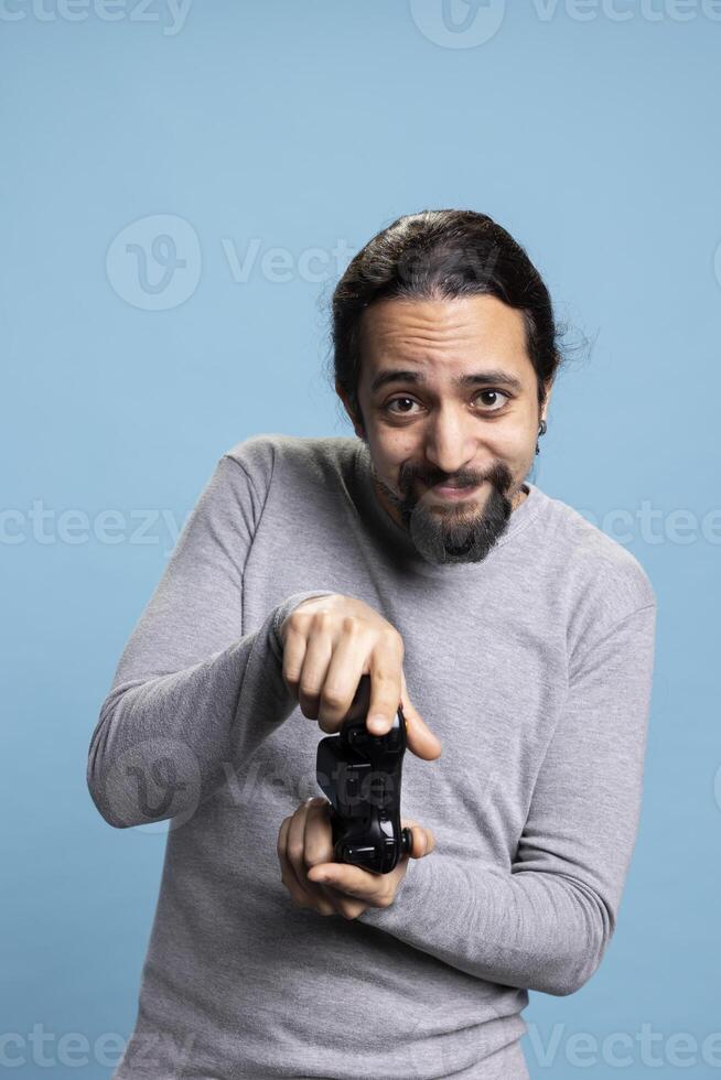 Middle eastern man playing games online on his console, attending an electronic cyber tournament against blue background. Young adult having fun with a shooter in studio, controller. photo