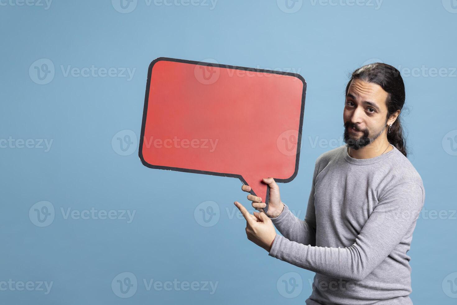 Young adult creating an advertisement with a speech bubble in studio, holding the empty red cardboard sign for a commercial against blue background. Male model uses carton copy space. photo