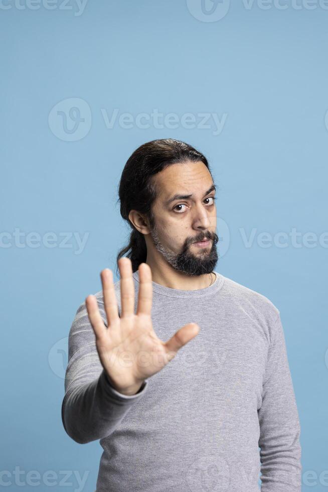 Young serious person expressing rejection with his palm raised, showing a stop sign against blue background. Male adult with a beard shows his refusal, irritated displeased reaction. photo