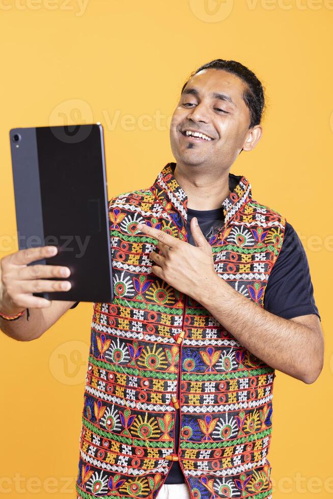 Happy indian man smiling, using tablet to take selfies from all angles. Vain social media user taking photos using device selfie camera, feeling thrilled, studio background