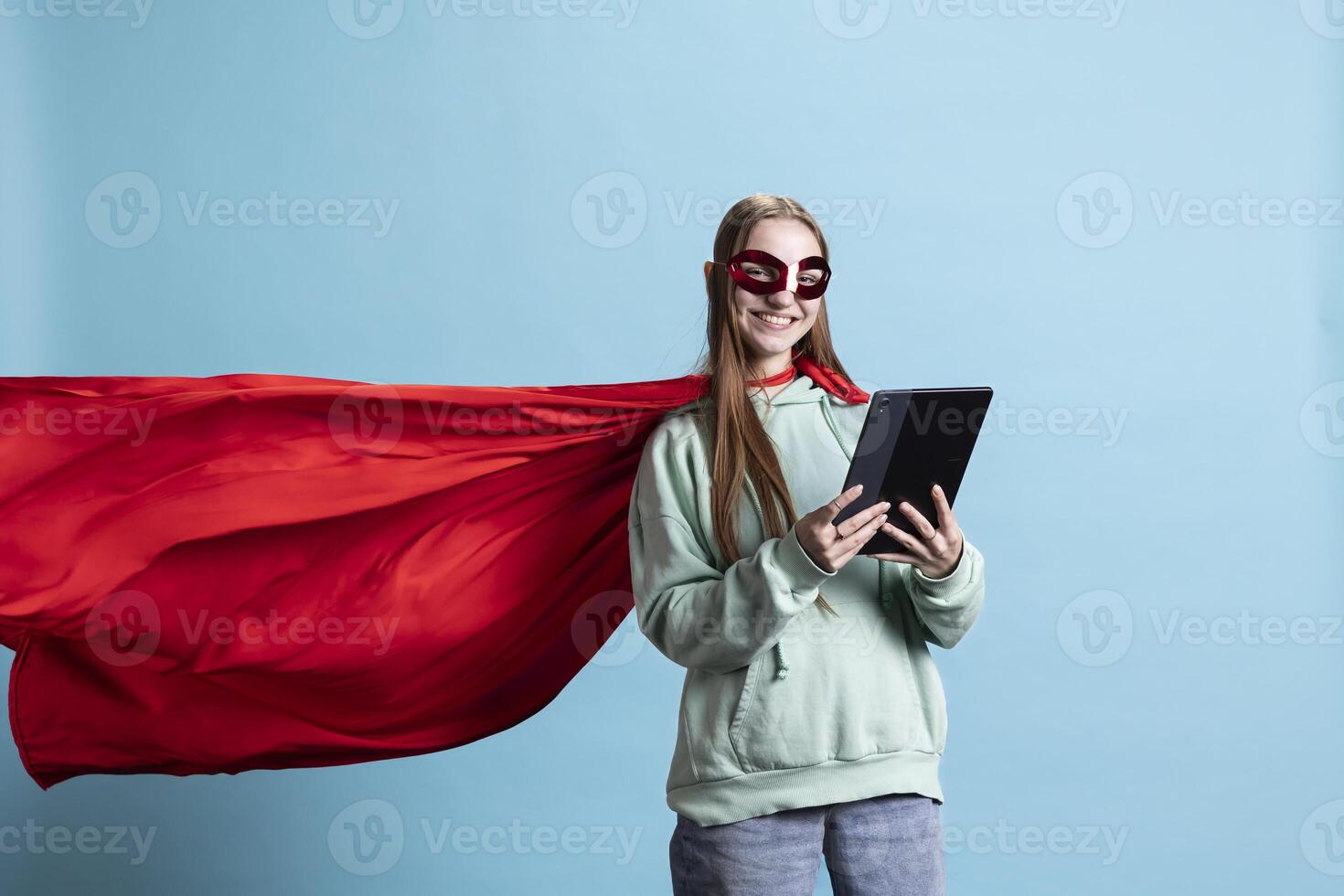 Joyous girl wearing superhero costume and mask for Halloween, using tablet, studio background. Portrait of upbeat person dressed as comic book hero hiding identity, holding device photo