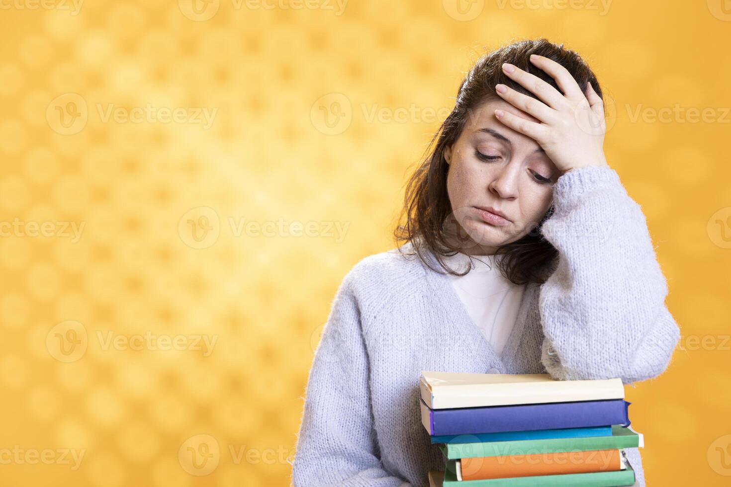 Woman with worried facial expression doing face palm from migraine pain, carrying books for school exam, studio background. Stressed student with textbooks suffering headache from too much homework photo
