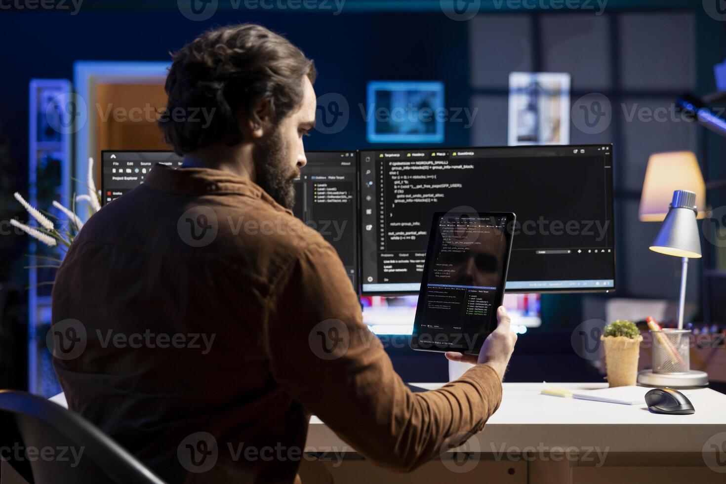 Programmer doing software debugging using tablet, checking glitches in lines of code displayed on computer monitors. Man working from home fixing coding issues using portable digital device photo