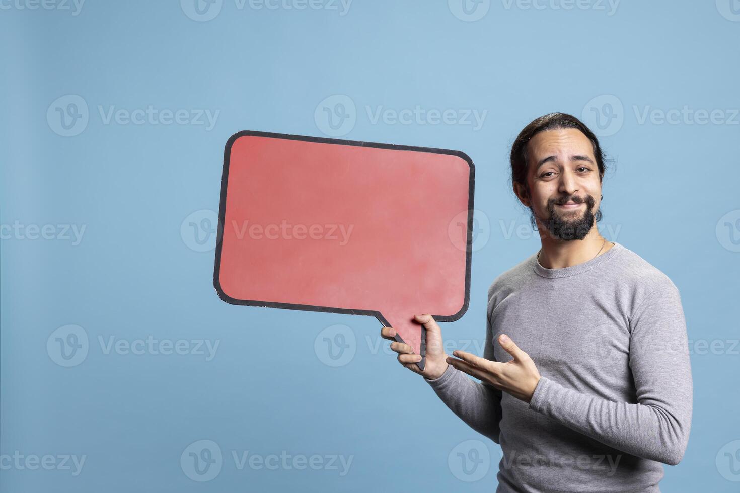 Young adult creating an advertisement with a speech bubble in studio, holding the empty red cardboard sign for a commercial against blue background. Male model uses carton copy space. photo