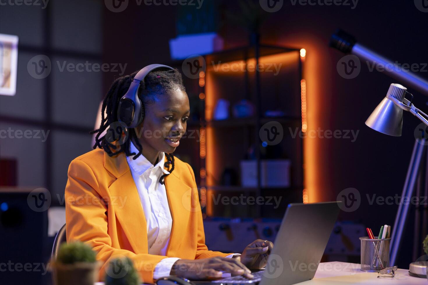 Remote worker listening music, analyzing data on laptop at desk, compiling analytical reports. Woman in home office wearing headphones, using notebook device, typing information photo