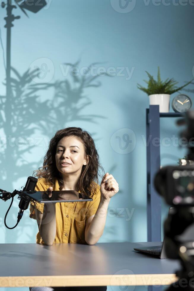 Upbeat tech engineer presenting tablet in entry level low budget price range, filming from dimly lit home studio. Jolly tech content creator reviewing digital device in front of online audience photo