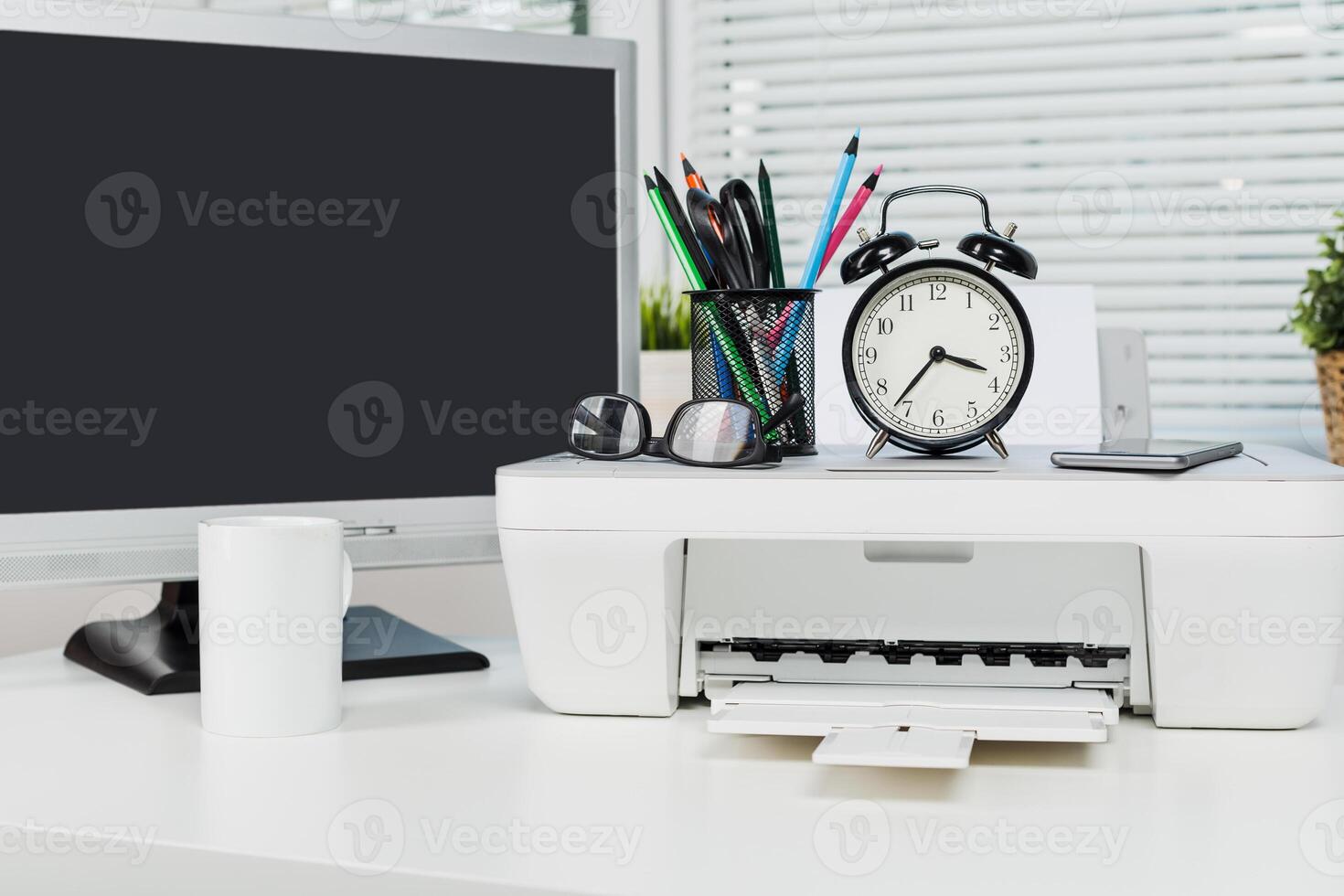 Office workspace featuring printer, clock, stationery, and computer set up during daytime hours photo