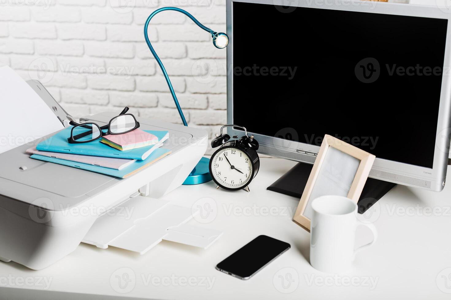 Workspace setup with printer, computer monitor, clock, and accessories on a desk during daylight photo