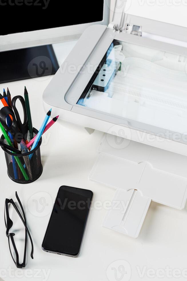 Office workspace with a printer, smartphone, stationery, and glasses on a desk during the day photo