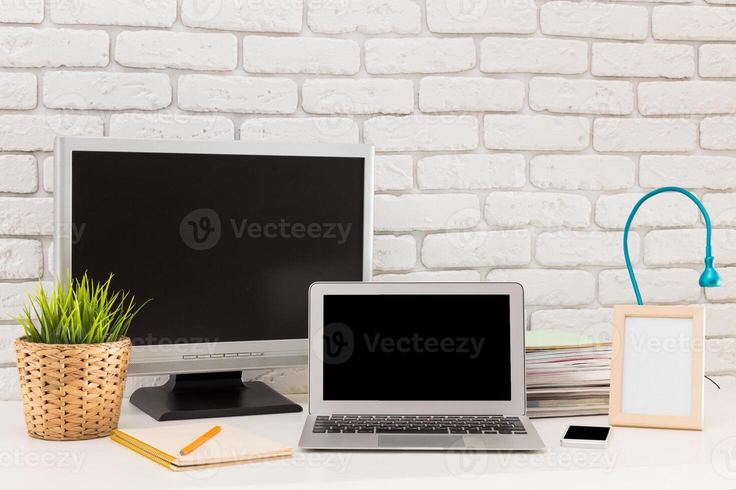 Workstation setup with a laptop, monitor, and stationery on a desk against a white brick wall in a well-lit space photo