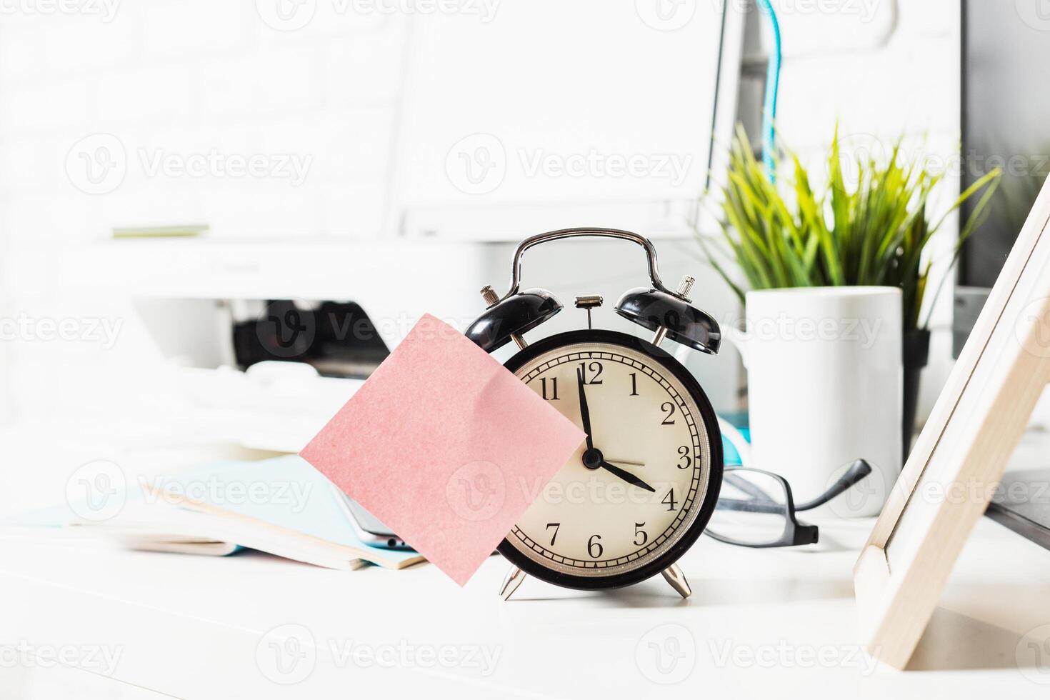 Clock on desk with sticky note and green plant during daytime work session photo