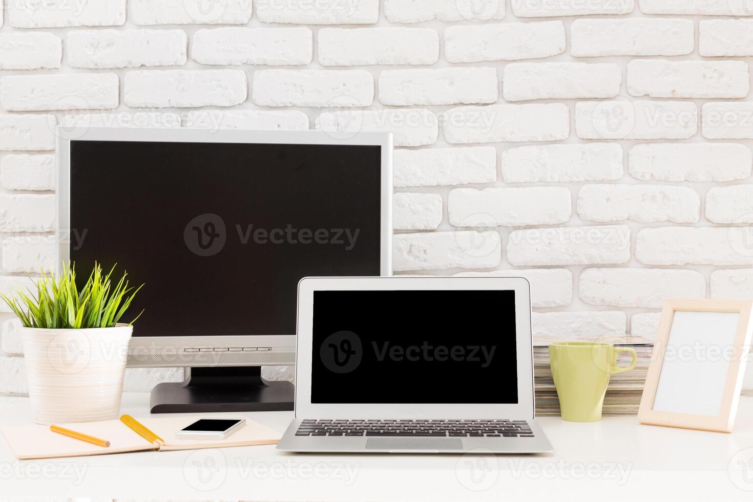 Modern workspace featuring laptops, plants, and stationery set against a brick wall backdrop during daylight photo