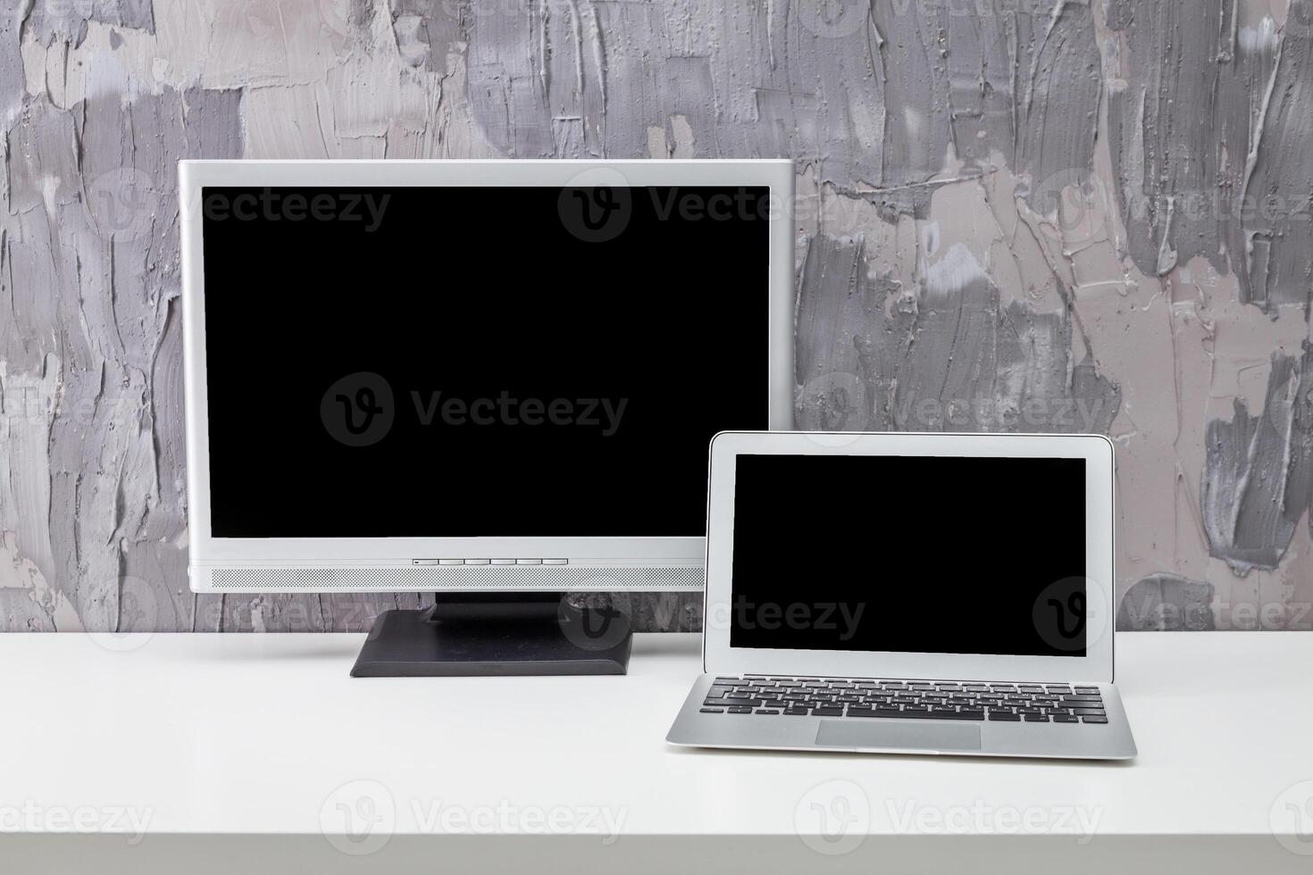 Computer setup with desktop monitor and laptop on a minimalist white table against a textured gray wall photo