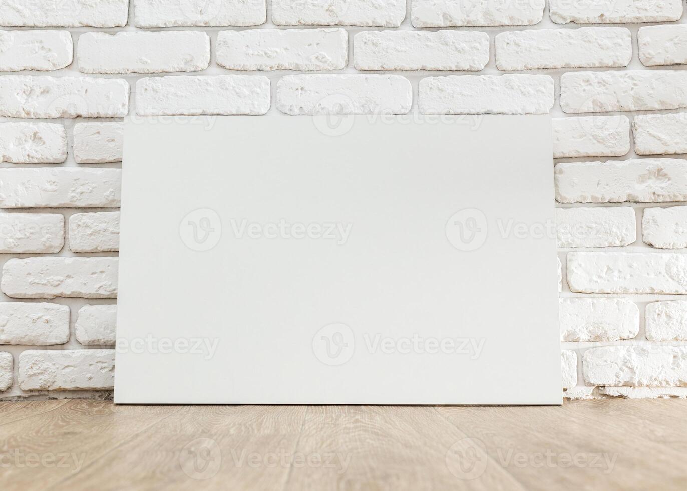 Blank white canvas leaning against a textured white brick wall beside light wooden flooring in a well-lit studio photo
