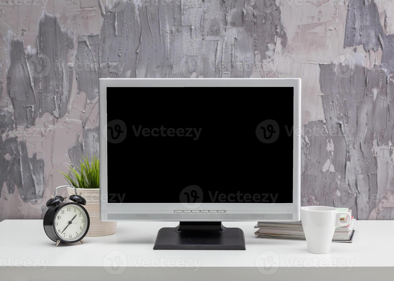 Modern workspace setup featuring a blank monitor, clock, plant, and coffee cup on a desk with textured wall background photo