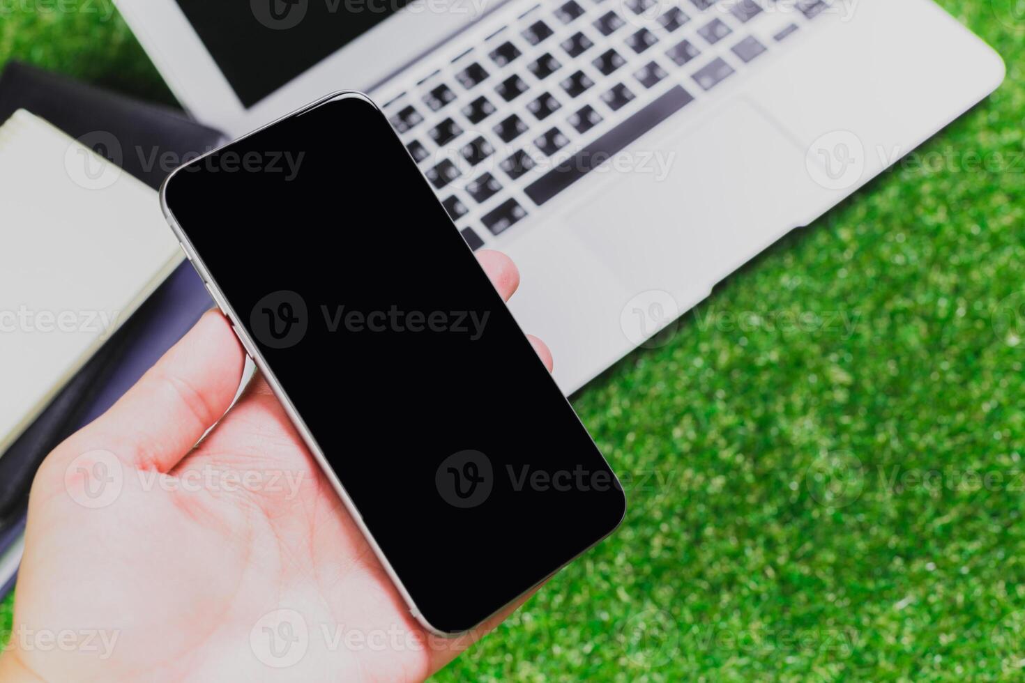 Hand holding a smartphone near a laptop on a grassy surface during a bright day photo