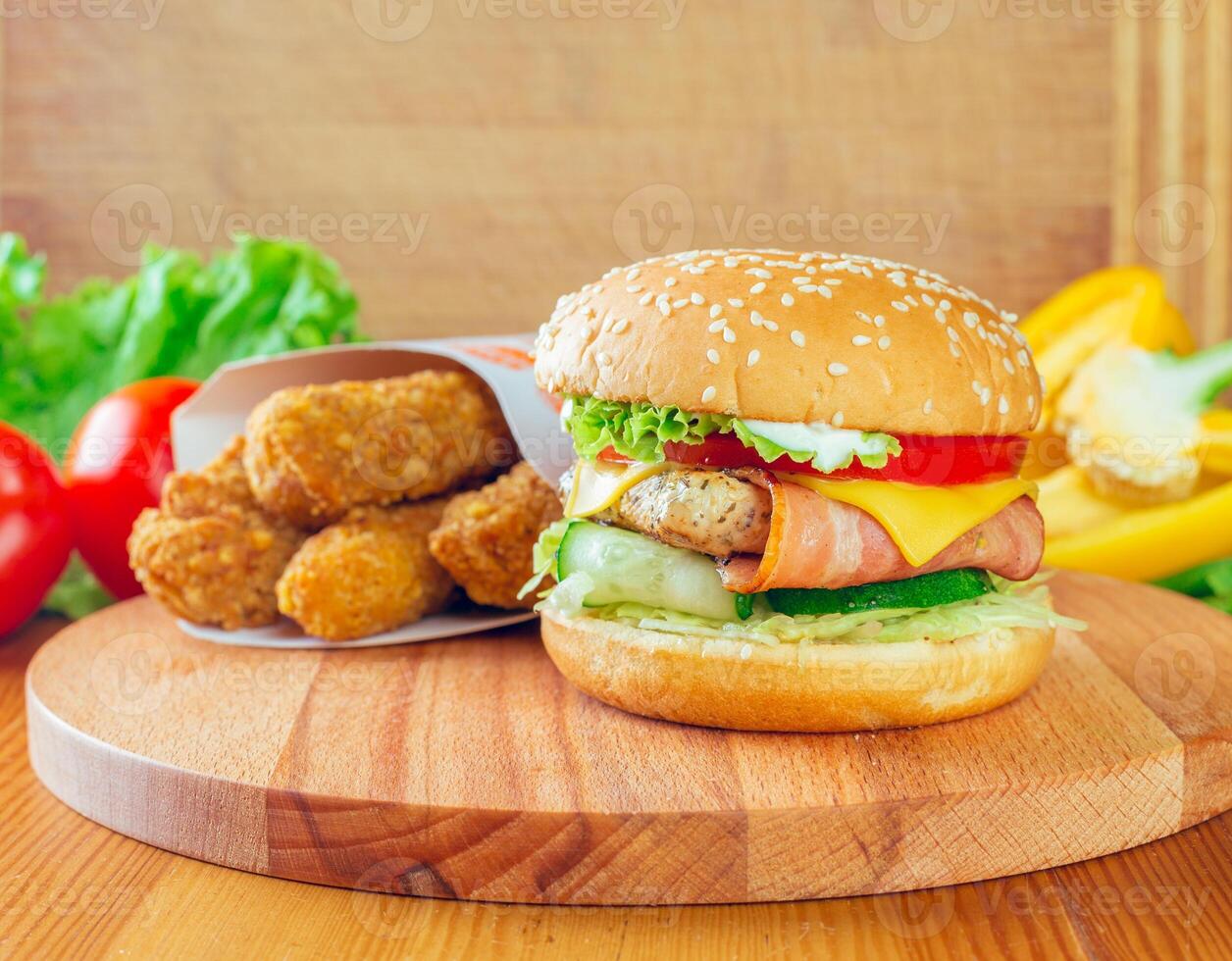Delicious burger with cheese, vegetables, and side of crispy chicken nuggets on wooden board photo