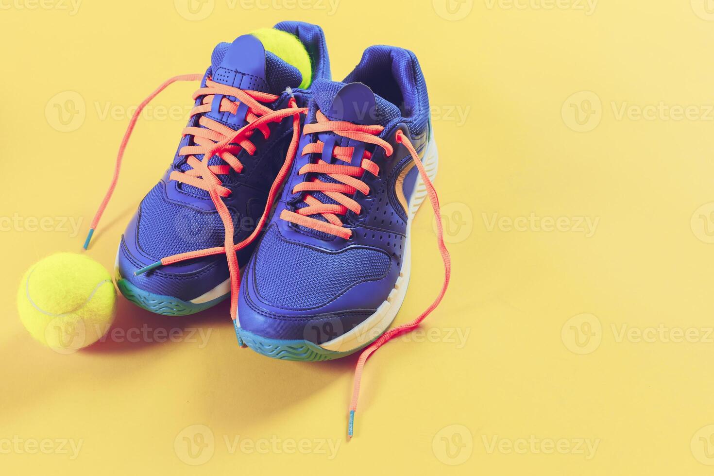 Bright blue athletic shoes with orange laces next to a tennis ball on a vibrant yellow background photo