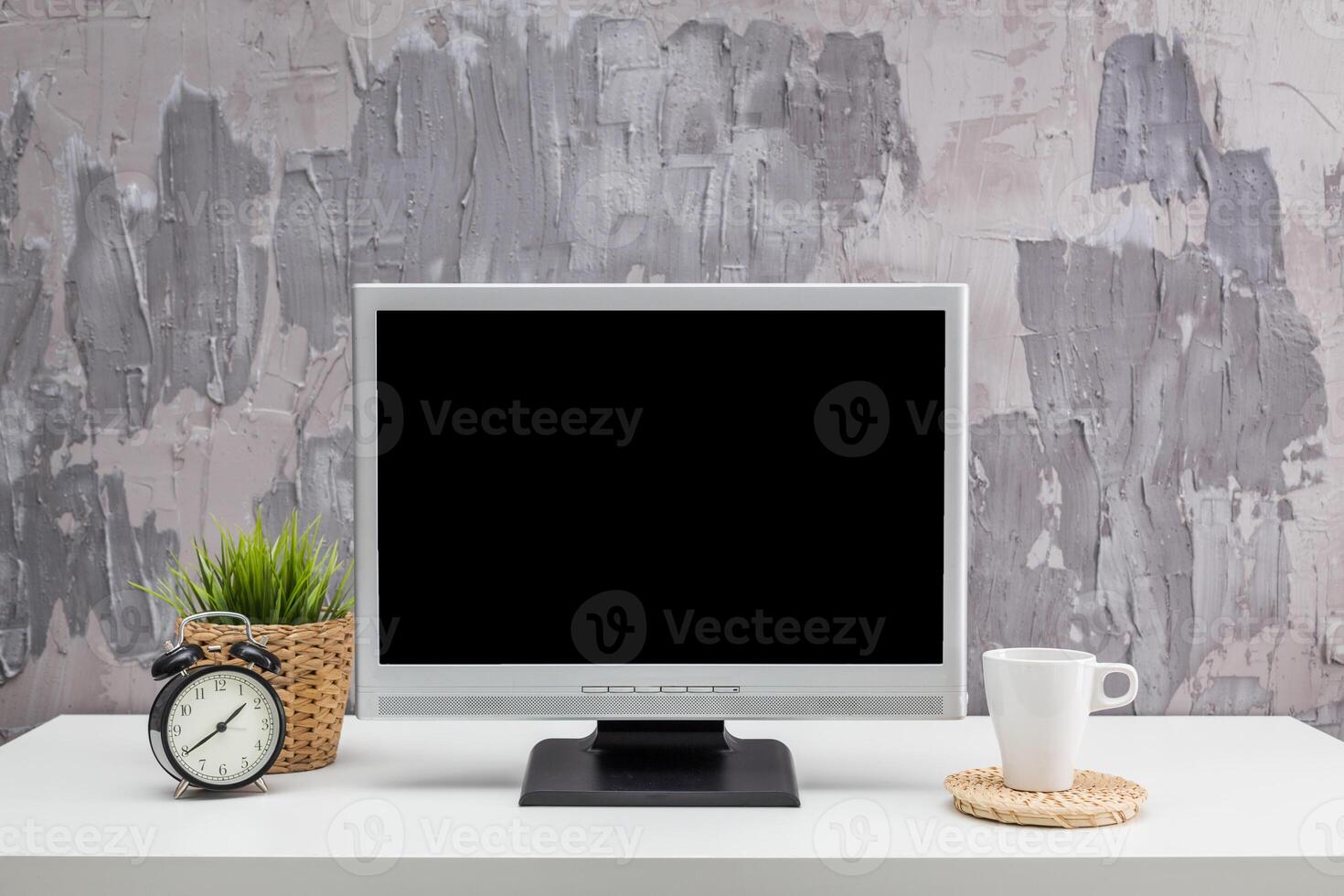 Desk setup featuring a blank computer monitor, clock, plant, and cup in a modern workspace with textured wall photo