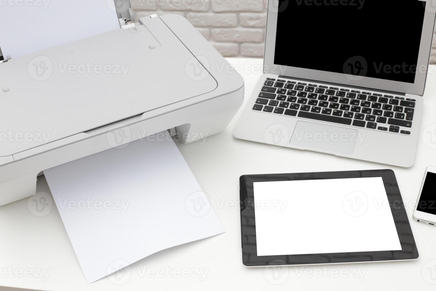 Printer in an office setting with a laptop, tablet, and smartphone on a desk during the day photo