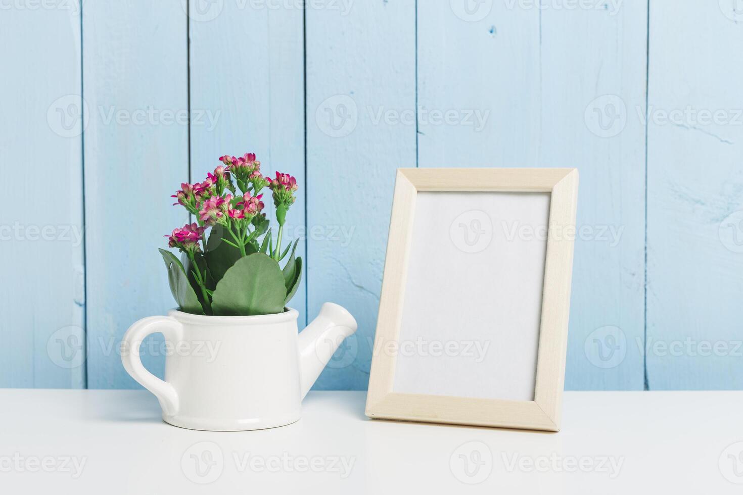 Colorful flower arrangement in a watering can beside a blank picture frame on a white table against a blue wooden background photo