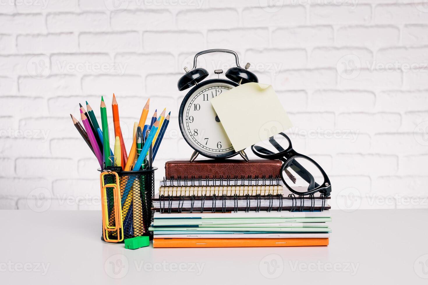 Organized workspace featuring stationery, an alarm clock, and colorful pencils on a white brick backdrop in a bright environment photo