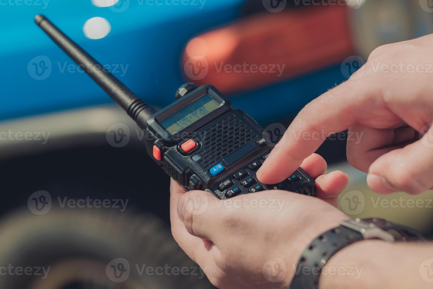 Individual operates handheld radio while standing next to blue vehicle in an outdoor setting during daytime photo
