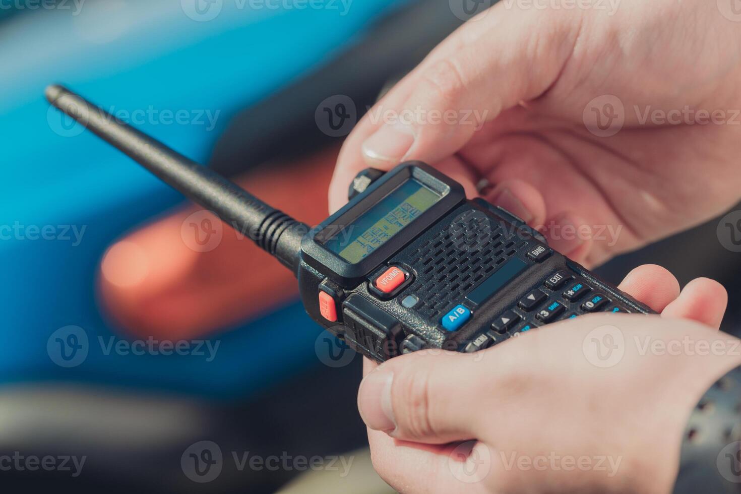 Hands hold a two-way radio while a blue vehicle is in the background during a sunny day photo