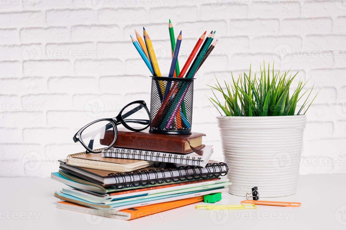 Workspace arrangement with colorful pencils, notebooks, and glasses on a white table against a brick wall backdrop photo