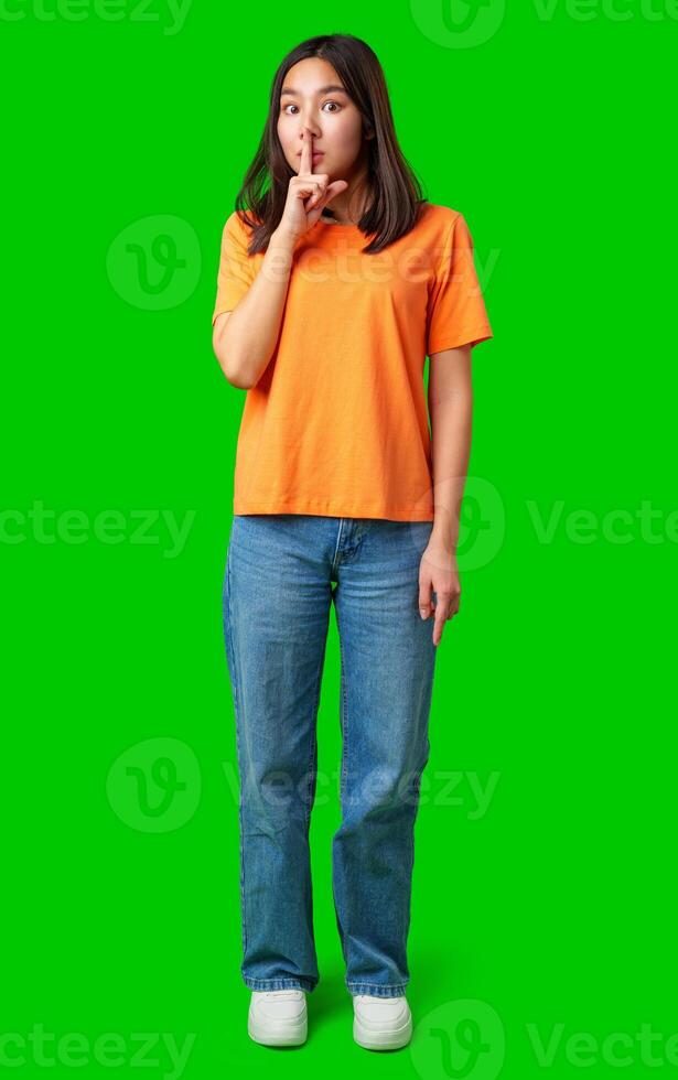 Young woman in orange shirt holding finger to lips on bright green background while signaling to keep quiet and maintain secrecy photo