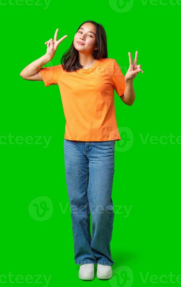 Cheerful young woman making peace signs with fingers while standing against a bright green background during daylight hours photo