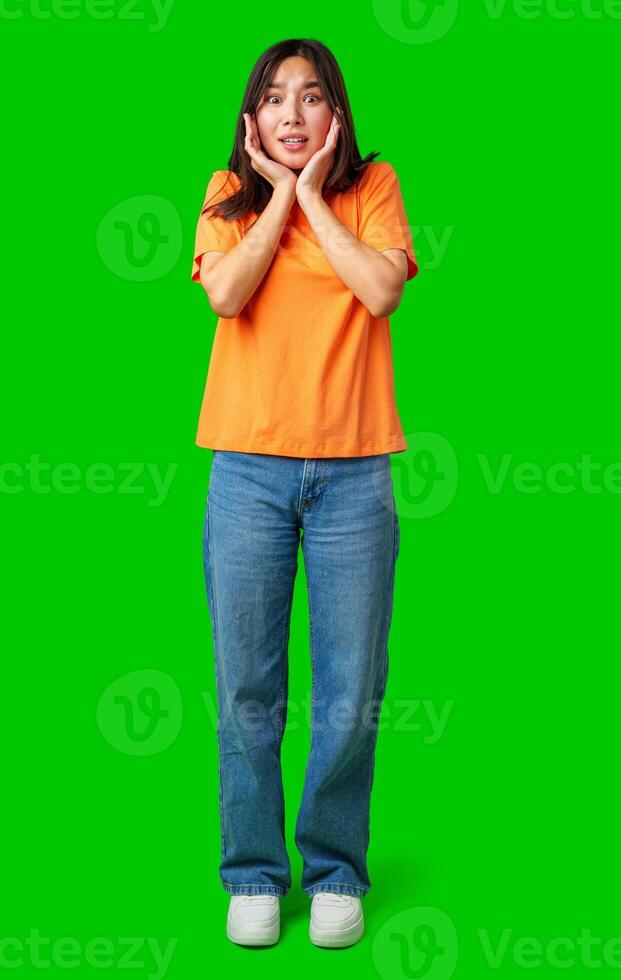 Young woman expresses surprise wearing a bright orange shirt and blue jeans against a green background photo