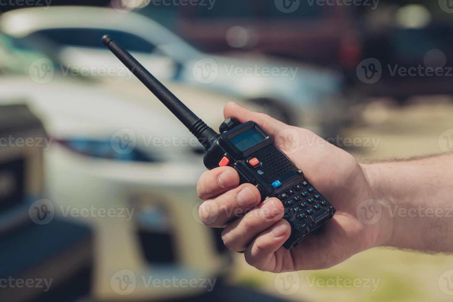 Hand holding a walkie-talkie with an extended antenna near vehicles in a parking area photo