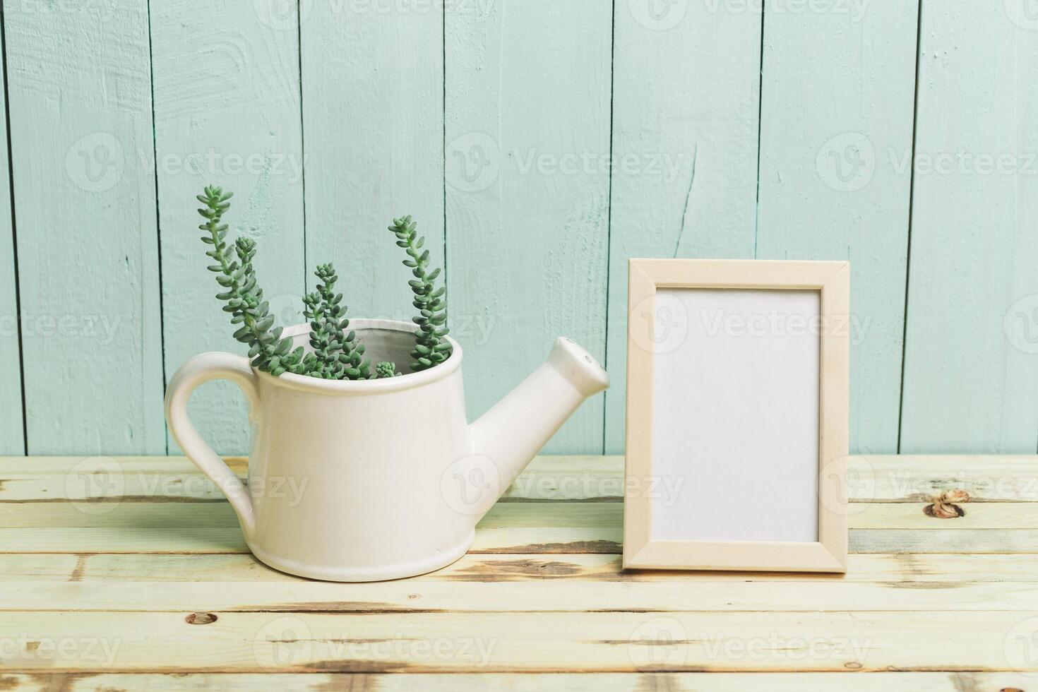 Succulent plant in a watering can next to a blank picture frame on a wooden table with a pastel background photo