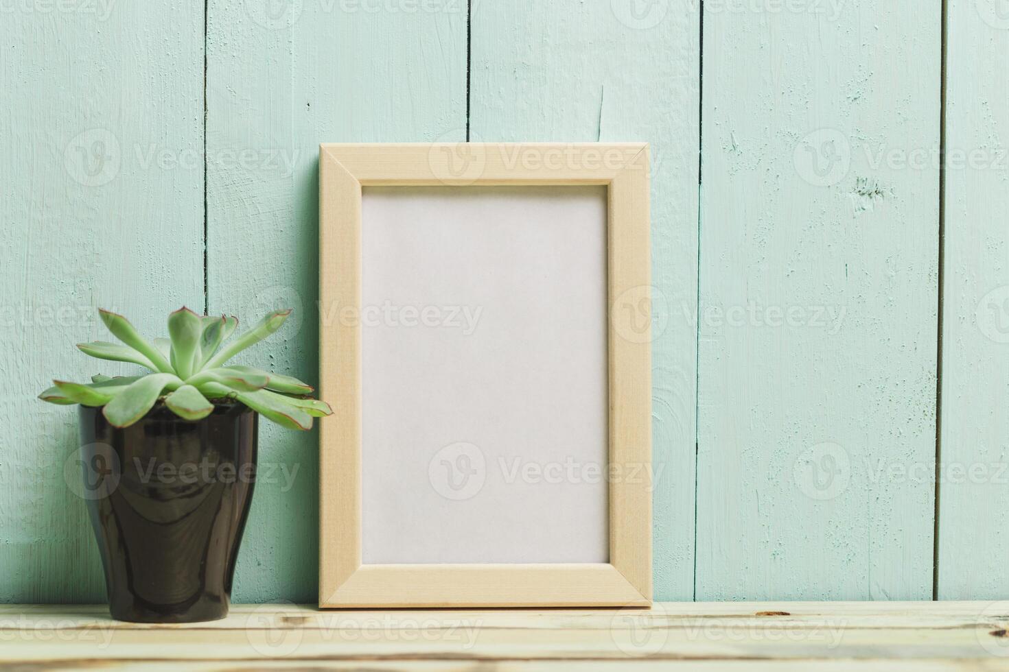 Decorative plant and empty picture frame on wooden table with teal background photo