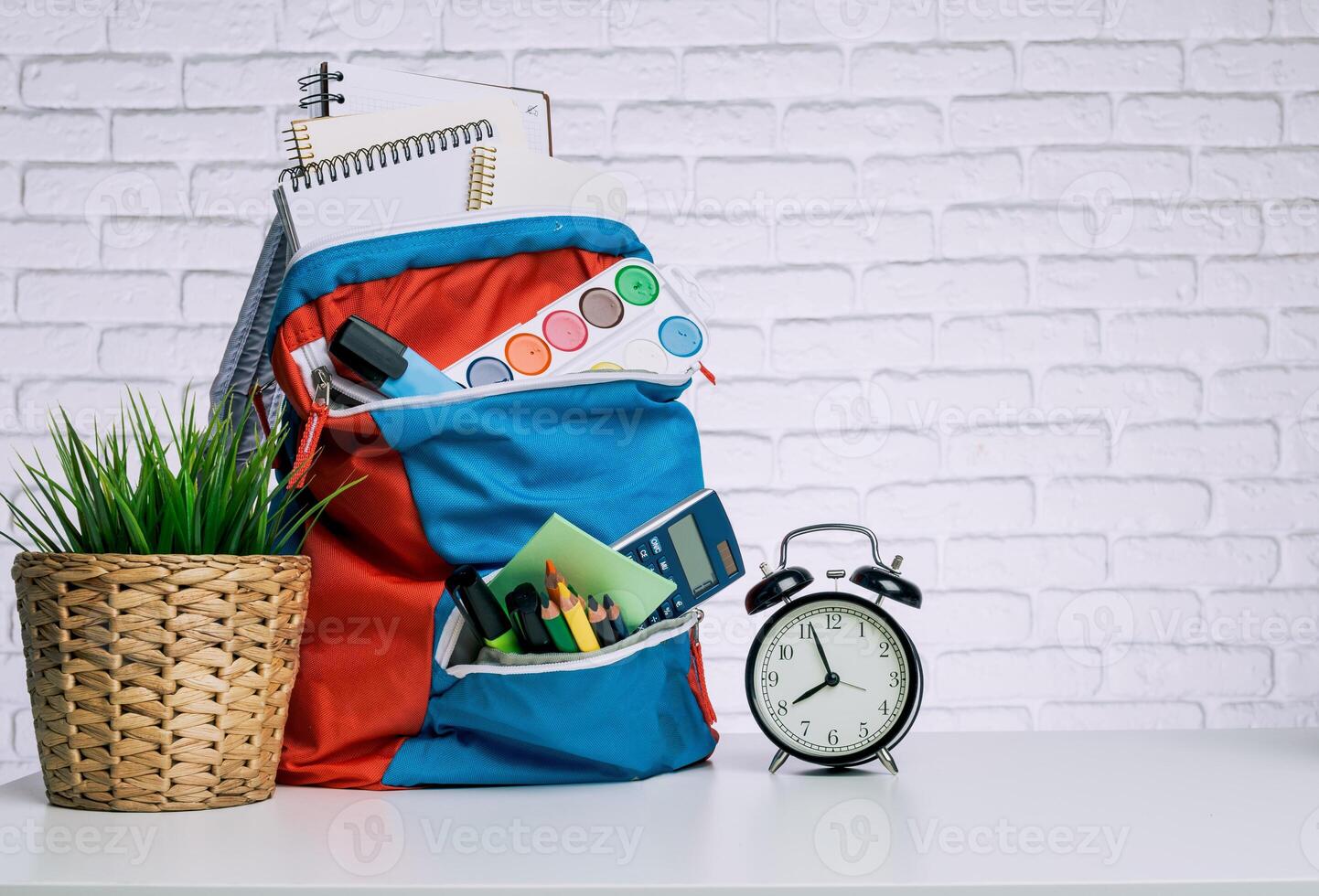 Colorful backpack on a table with school supplies, clock, and plant against a brick wall background photo