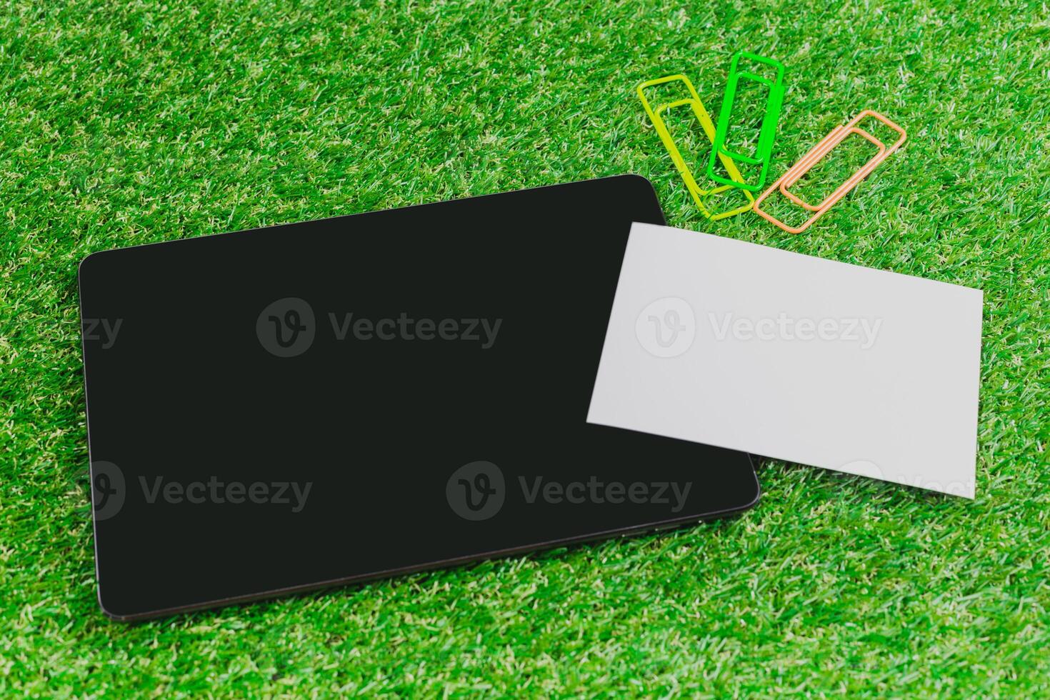 Workspace setup with tablet, blank card, and colorful paper clips on green grass background photo