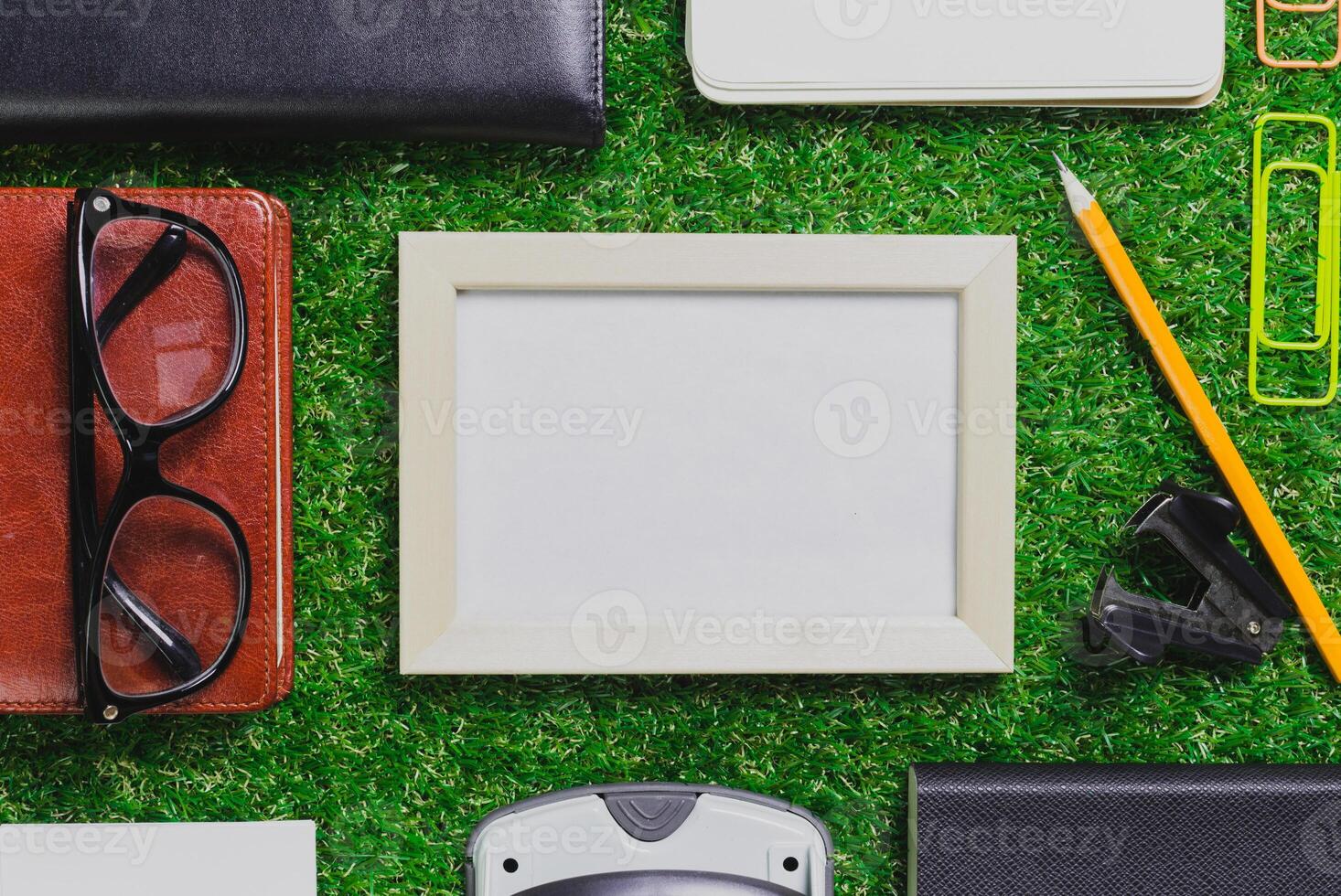 Collection of stationary items arranged on green grass with empty frame and office tools at midday photo