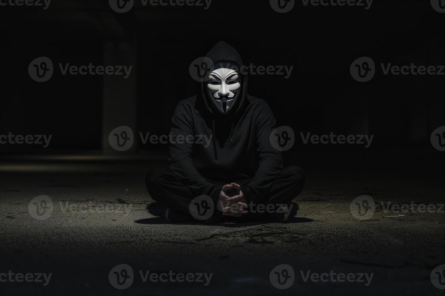 A man in a black hoodie with a fake smiley face mask on his face sits on the gro photo