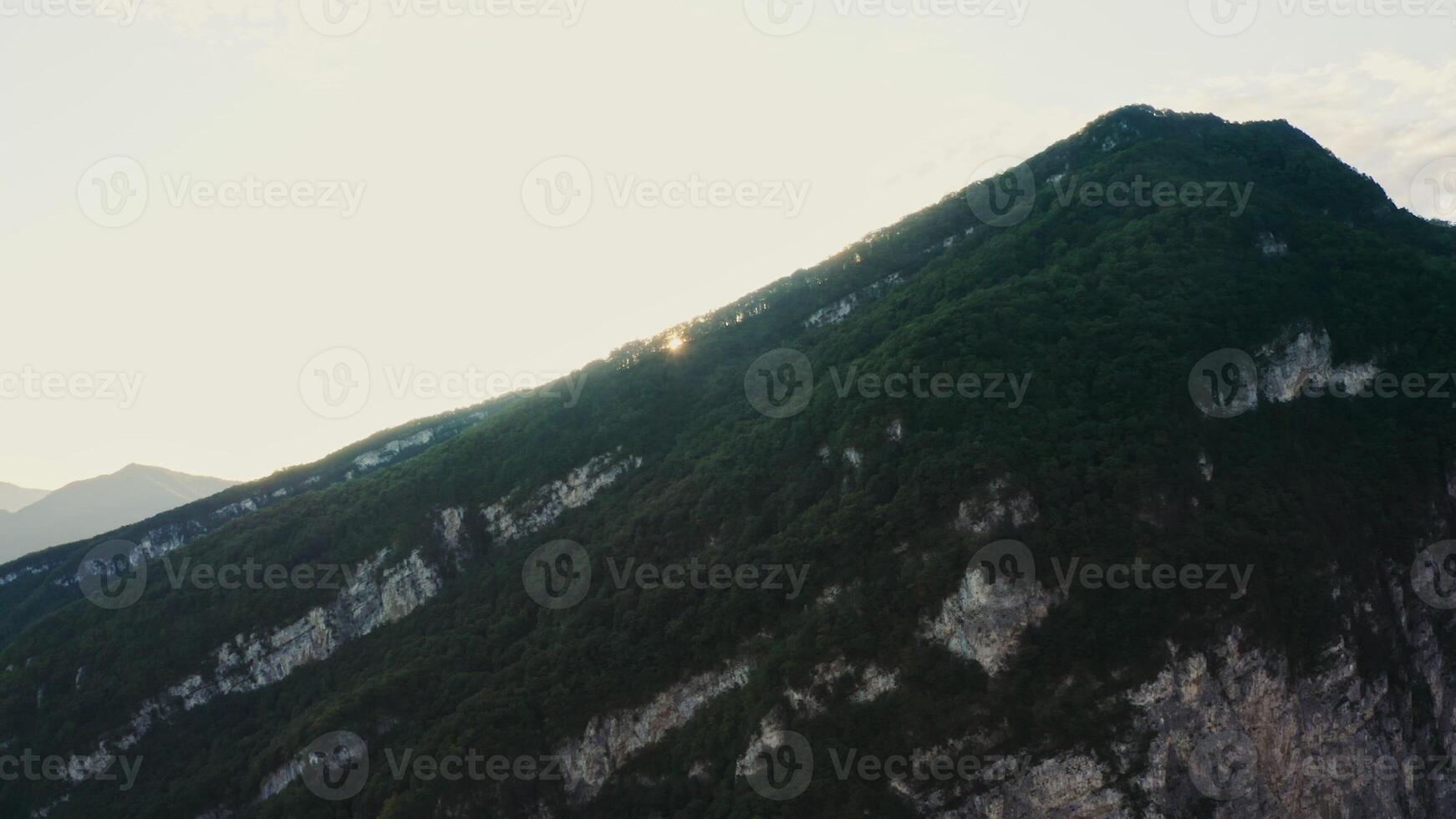 The sun appears from behind a rocky mountain slope overgrown with dense forest. Sunrise in the mountains, aerial view from a drone. Mountain peaks of the Caucasus photo
