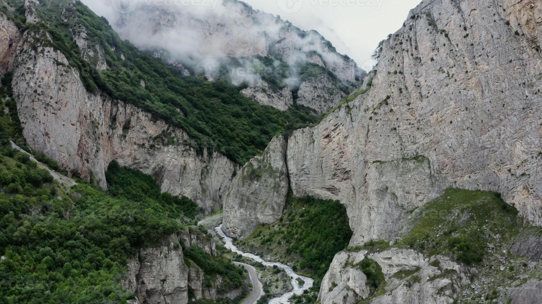 Vertical panning from a quadcopter. A narrow mountain road and a bubbling river in the lowlands between sheer rocky mountains covered with green vegetation and clouds photo