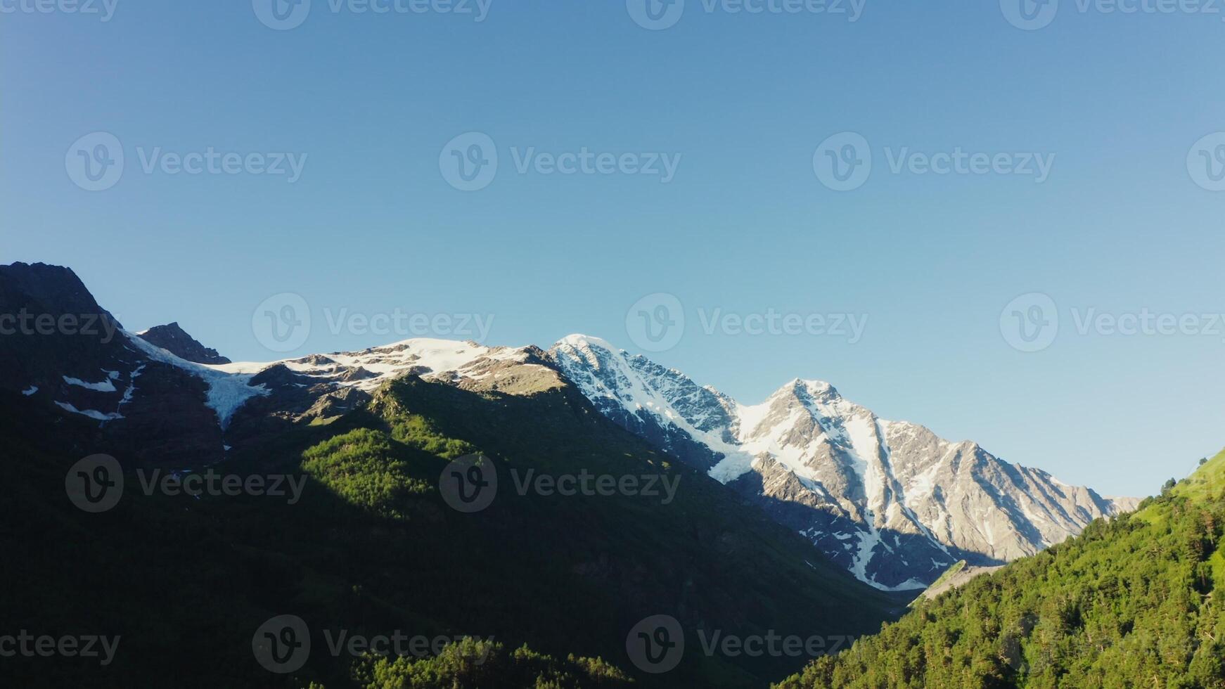 The snow-capped peak of Elbrus among the mountains covered with green forests. Mountain valley aerial view, shooting from a quadrocopter. Mountain peaks from drone photo