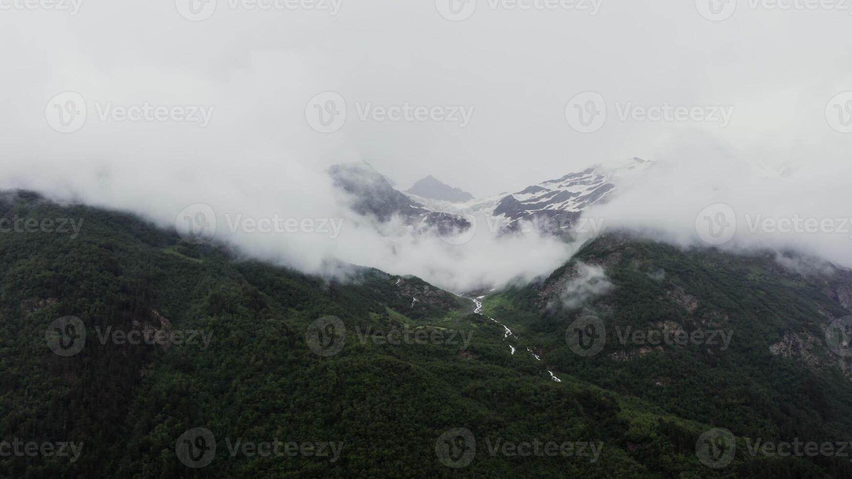 A bird's eye view of mountain valley with narrow, branched mountain river flowing between dense forest in lowland between the mountains, at the foot of rocky mountains covered with snow and clouds photo
