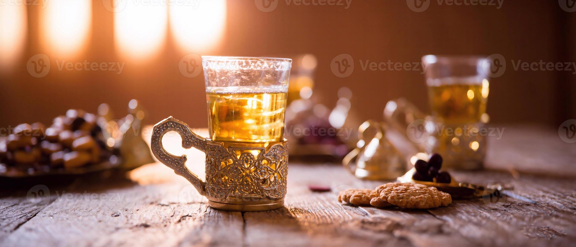 Wooden table set with a cup of tea in an ornate holder, a plate of dates, and a small bowl of dates. Soft, warm lighting and additional cups of tea. photo