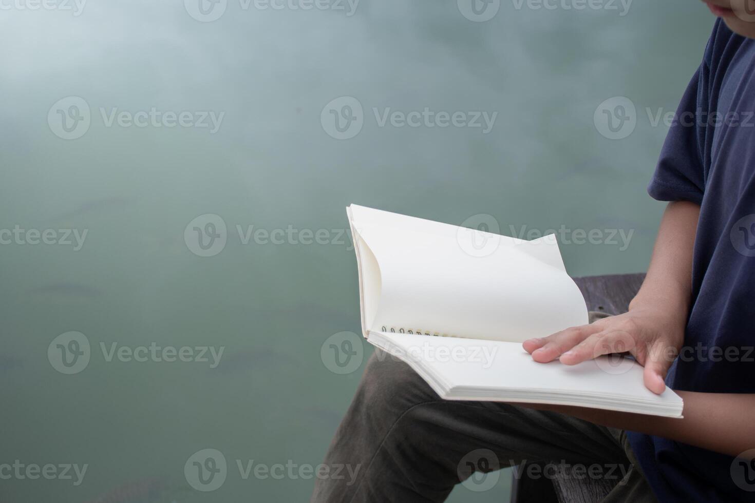 Boy opening blank book sitting on wooden bench at lakeside on water surface background,concept of education and work,blank notebook copy space for design,Hand opening blank book,boy holding notebook. photo