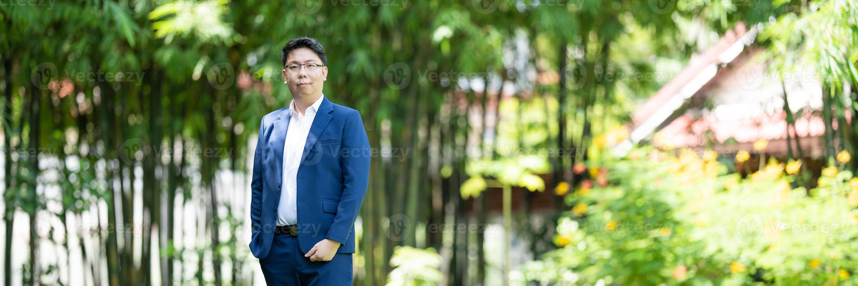 Asian handsome glasses man in a blue suit and the same color pants is looking at the camera, smart smiling and standing in the garden. photo