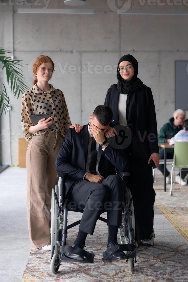 A diverse business team poses confidently, including a smiling director seated in a wheelchair, flanked by two professional female colleagues, one wearing a hijab and the other holding a tablet, in a photo